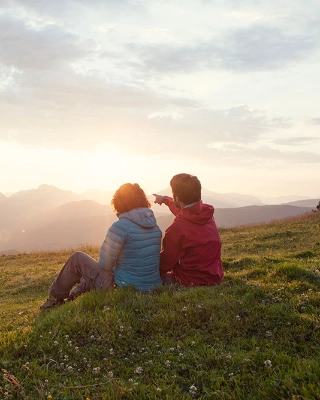 Ein Paar sitzt waehrend einer Wanderung auf der Wiese und blickt ueber das Tal.